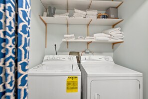 A laundry room with two front-loading washing machines, a shelf with folded towels, and a Verizon Internet Gateway box. - Blocks from the Ocean in a Convenient and Desirabl (Saint Simons Island)