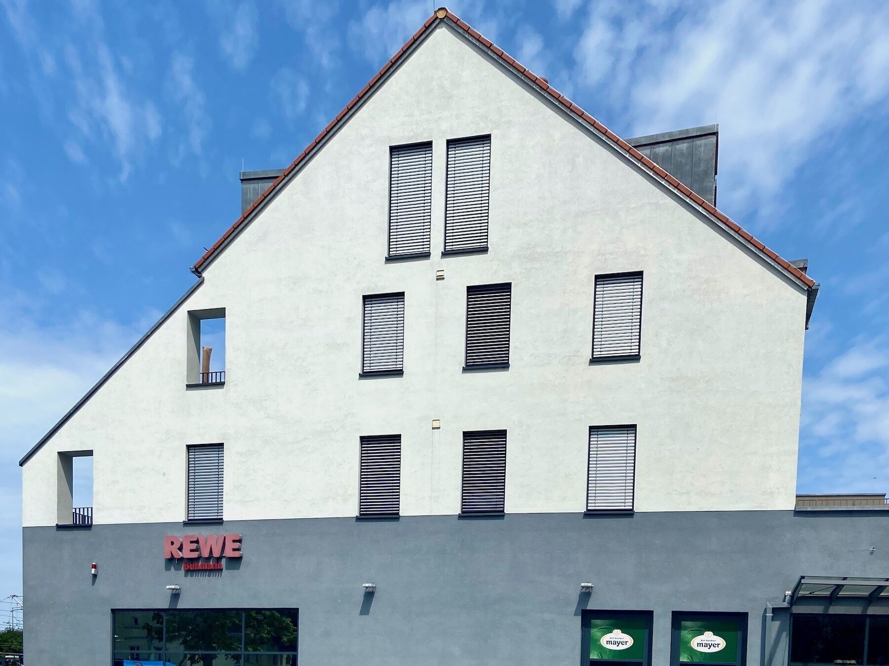 A modern building with a flat roof, multiple windows with shutters, and a sign for REWE.
