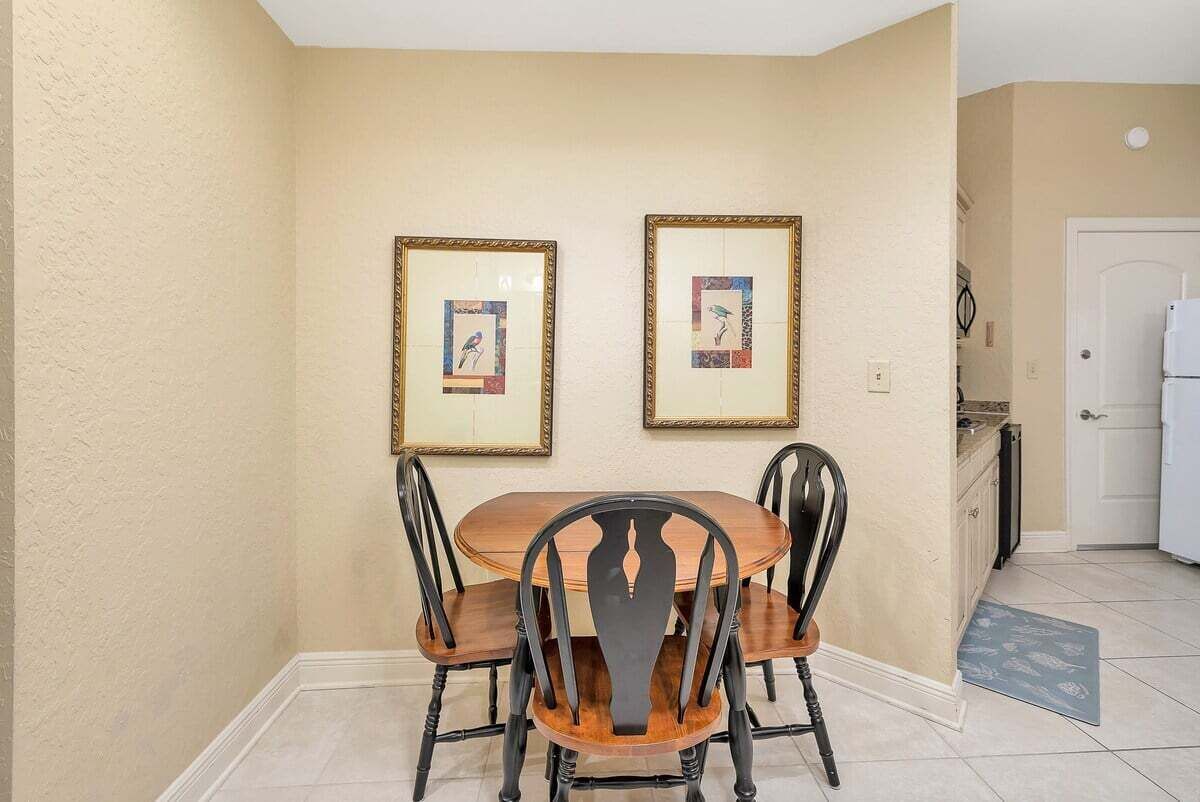 Un coin repas avec une table en bois et quatre chaises, des œuvres d’art encadrées au mur, et une cuisine en arrière-plan.