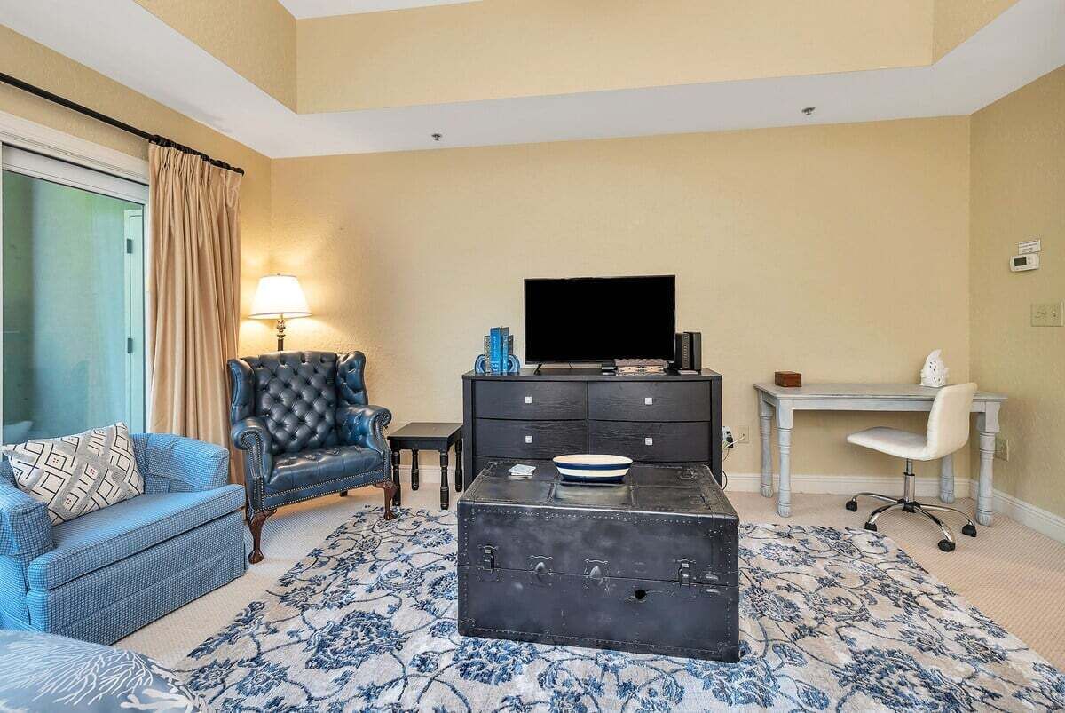 A living room with a blue sofa, a leather armchair, a dark chest of drawers, a flat-screen TV, a desk with a chair, and a patterned rug.