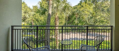 A balcony with two metal chairs and a small round table, overlooking a parking area and lush greenery.