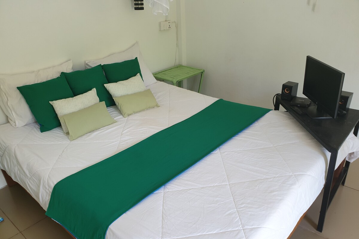 A neatly made bed with a green and white bedspread, a green nightstand with a lamp, and a desk with a computer and speakers.