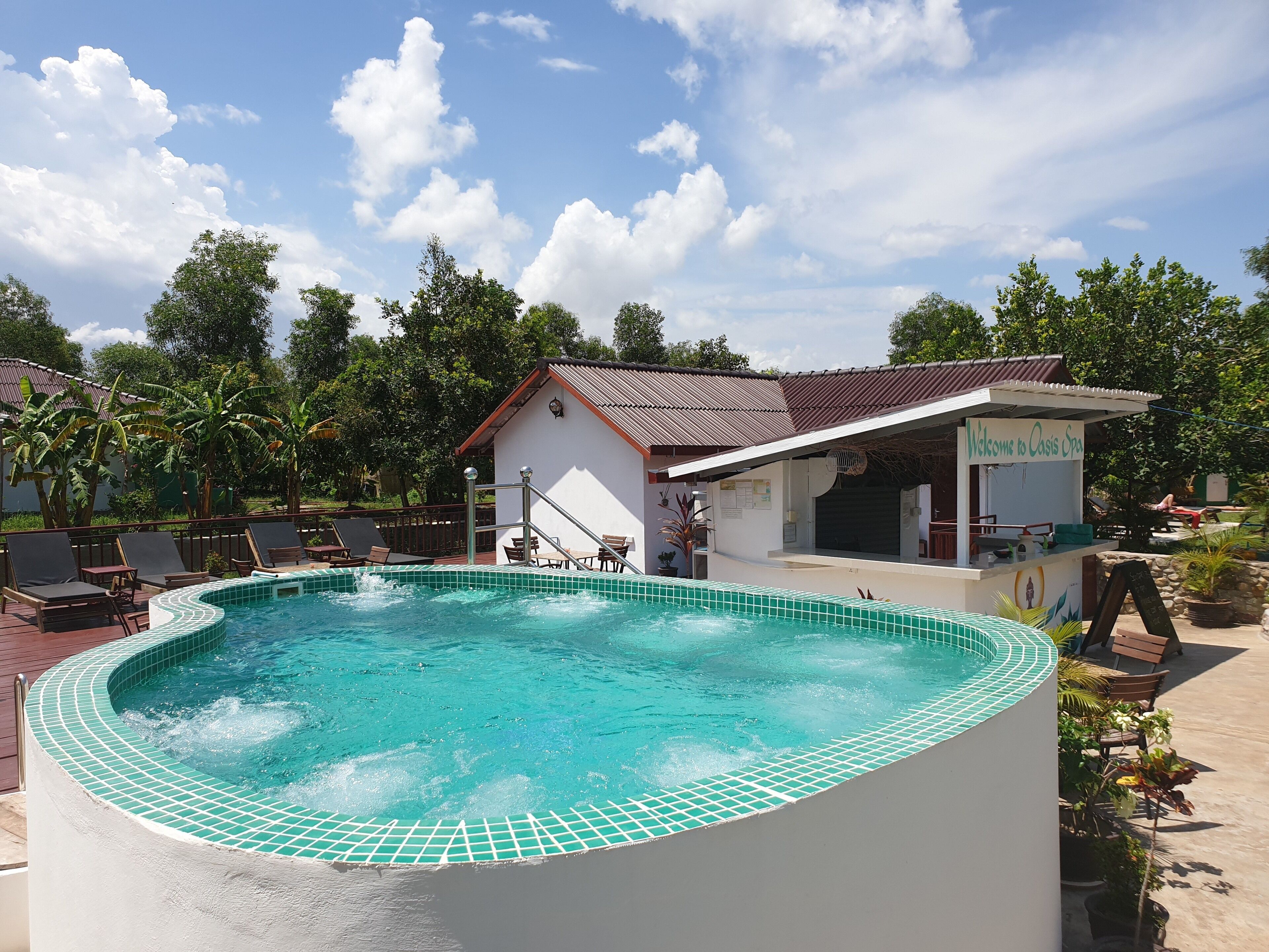 Una piscina con borde de azulejos, un pequeño edificio con un cartel que dice "Welcome to Oasis Spa", y un área circundante con sillas de descanso y plantas en macetas.