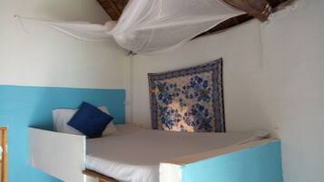 A single bed with a white mattress and blue pillows, a wooden headboard, a blue accent wall, a patterned tapestry, and a mosquito net above the bed.