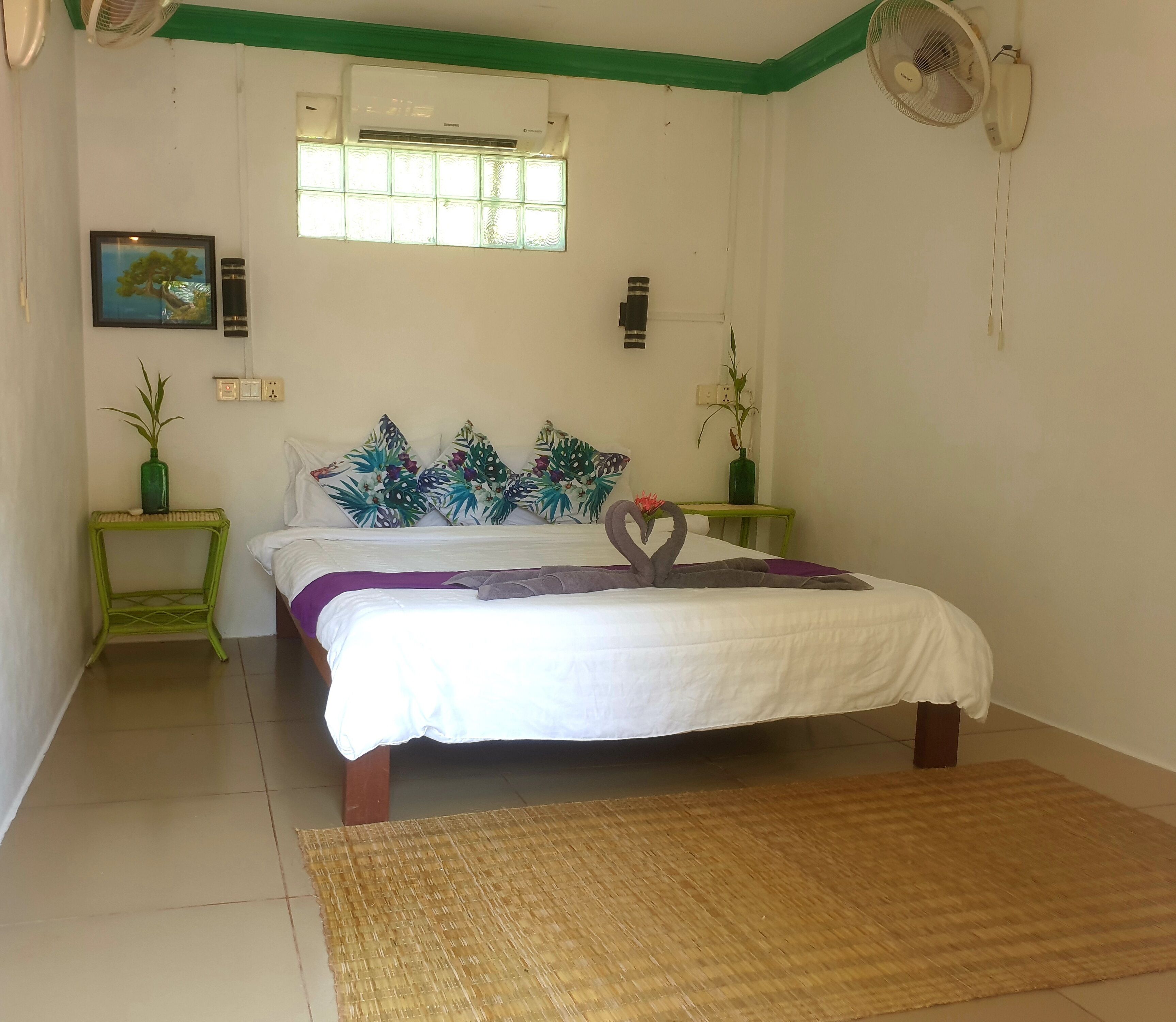 Un dormitorio ordenado con una cama, una ventana con persianas, un aire acondicionado de pared, una mesita con una planta y un ventilador de pared.