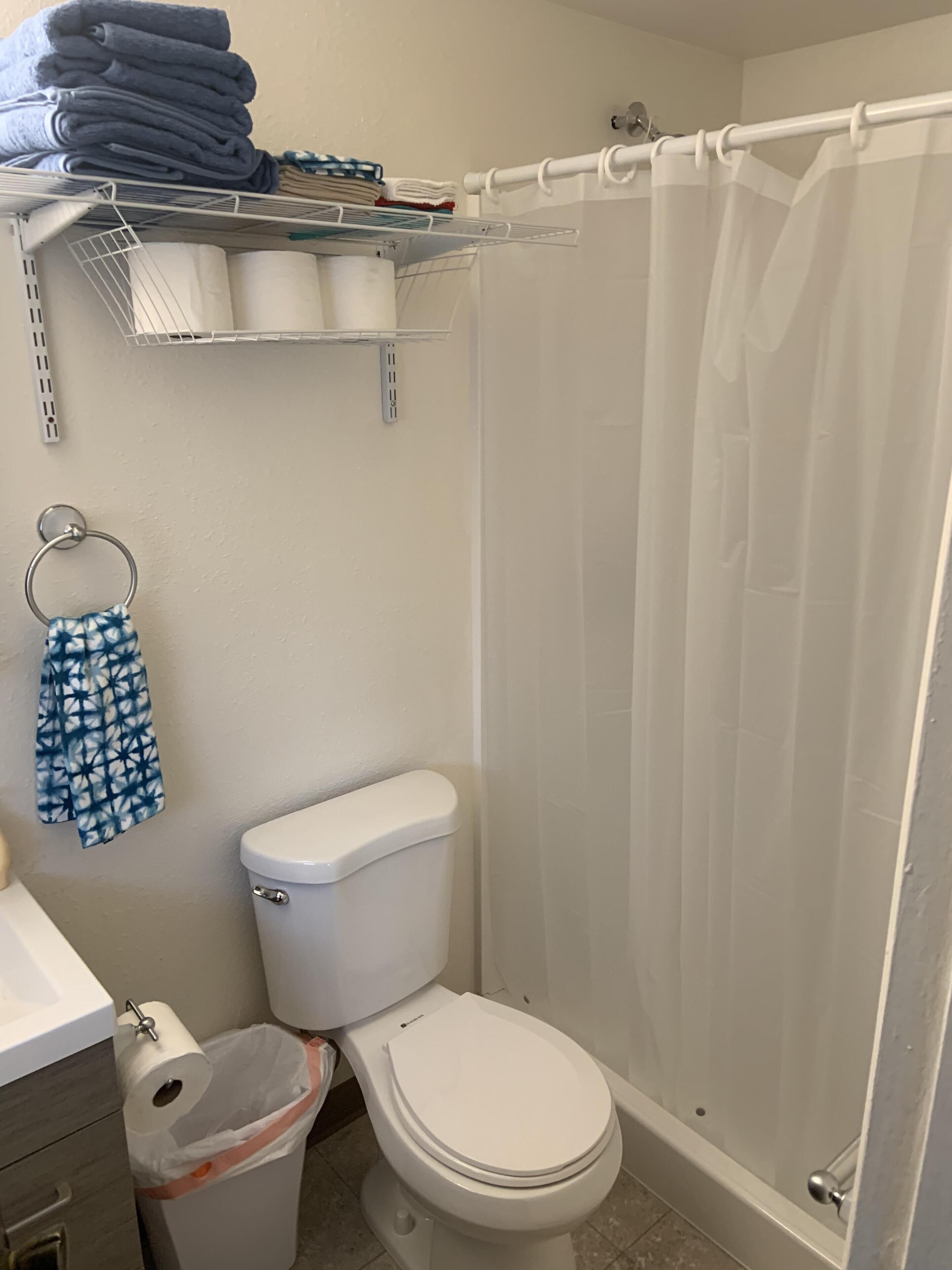 A bathroom with a toilet, a shower curtain, a towel rack with folded towels, and a trash bin.