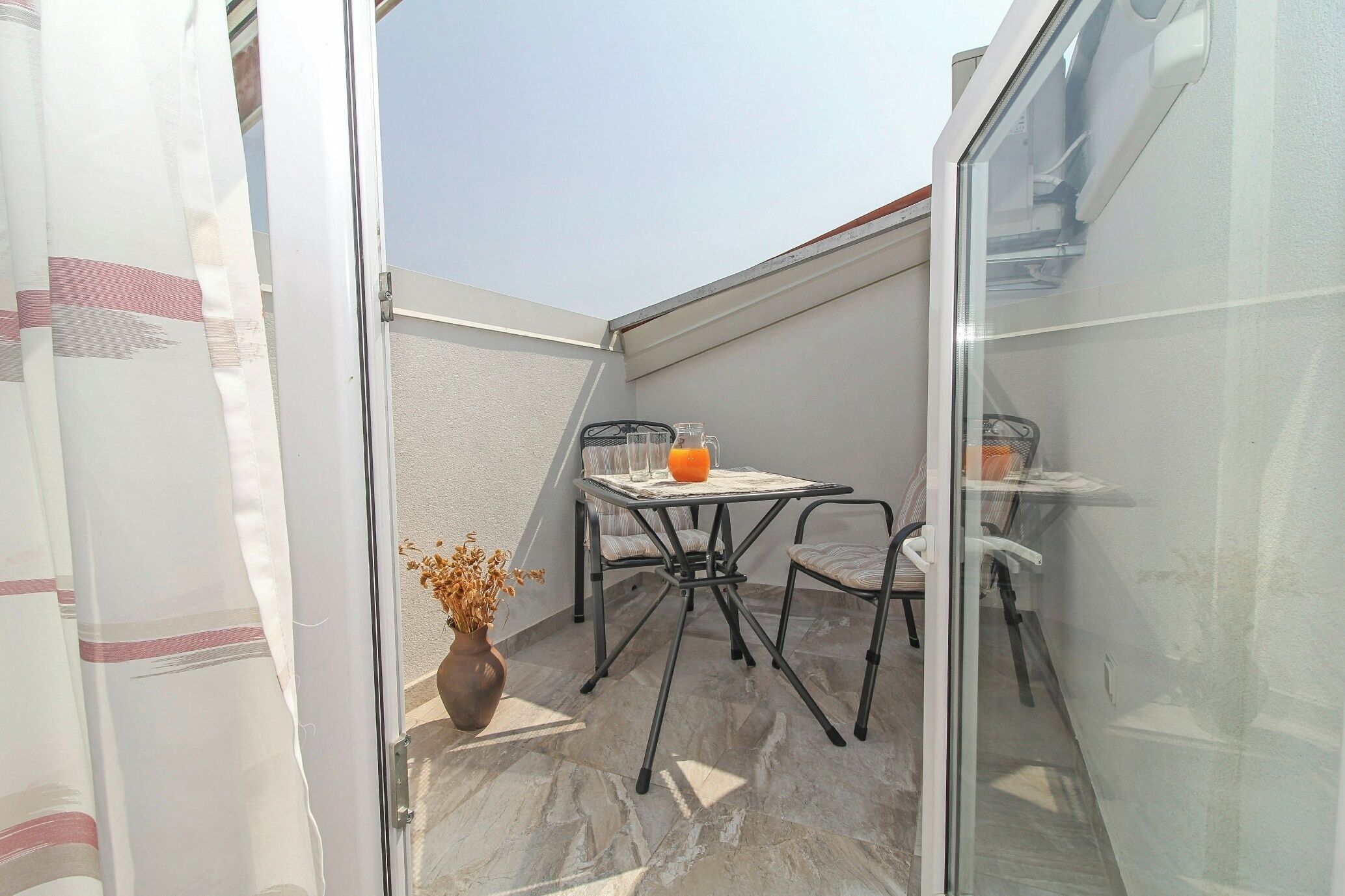 Un petit balcon extérieur avec une table et deux chaises, une plante en pot et une porte vitrée.