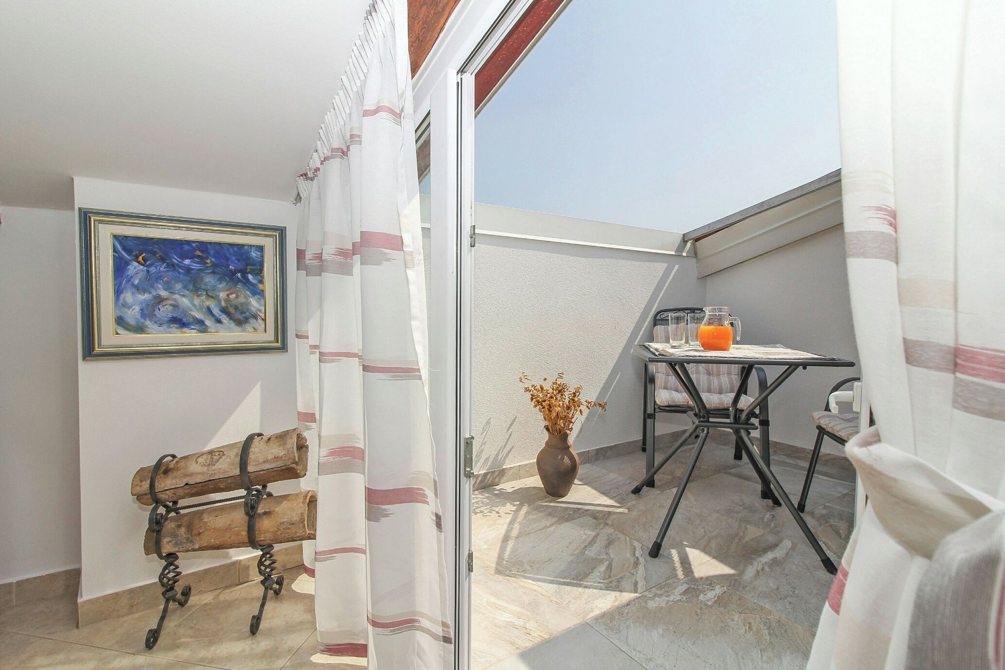 Un balcon avec une table, un banc, un vase contenant des fleurs séchées et un tableau accroché au mur.