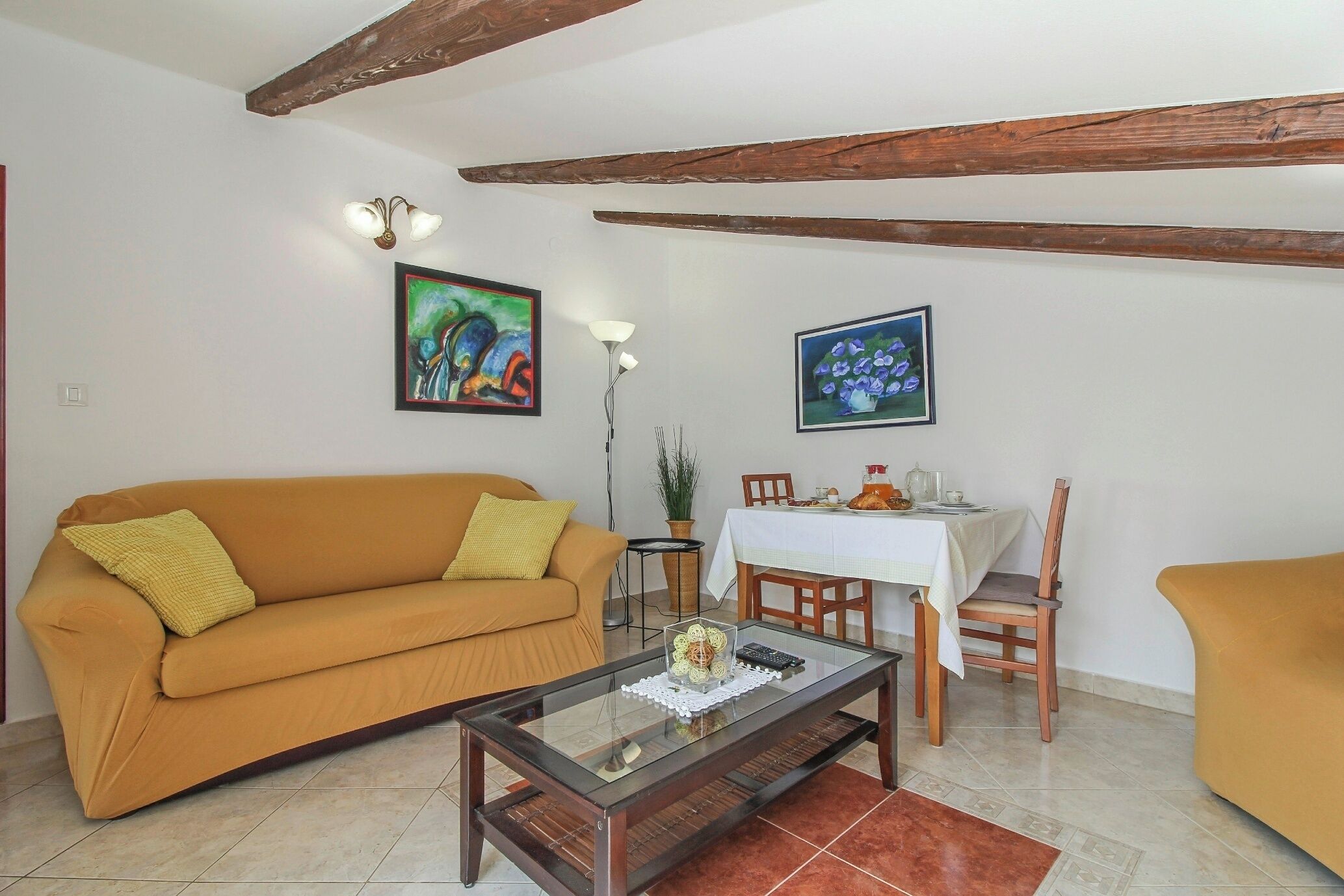 A living room with a sofa, coffee table, dining table, and chairs. There are paintings on the wall and a potted plant.