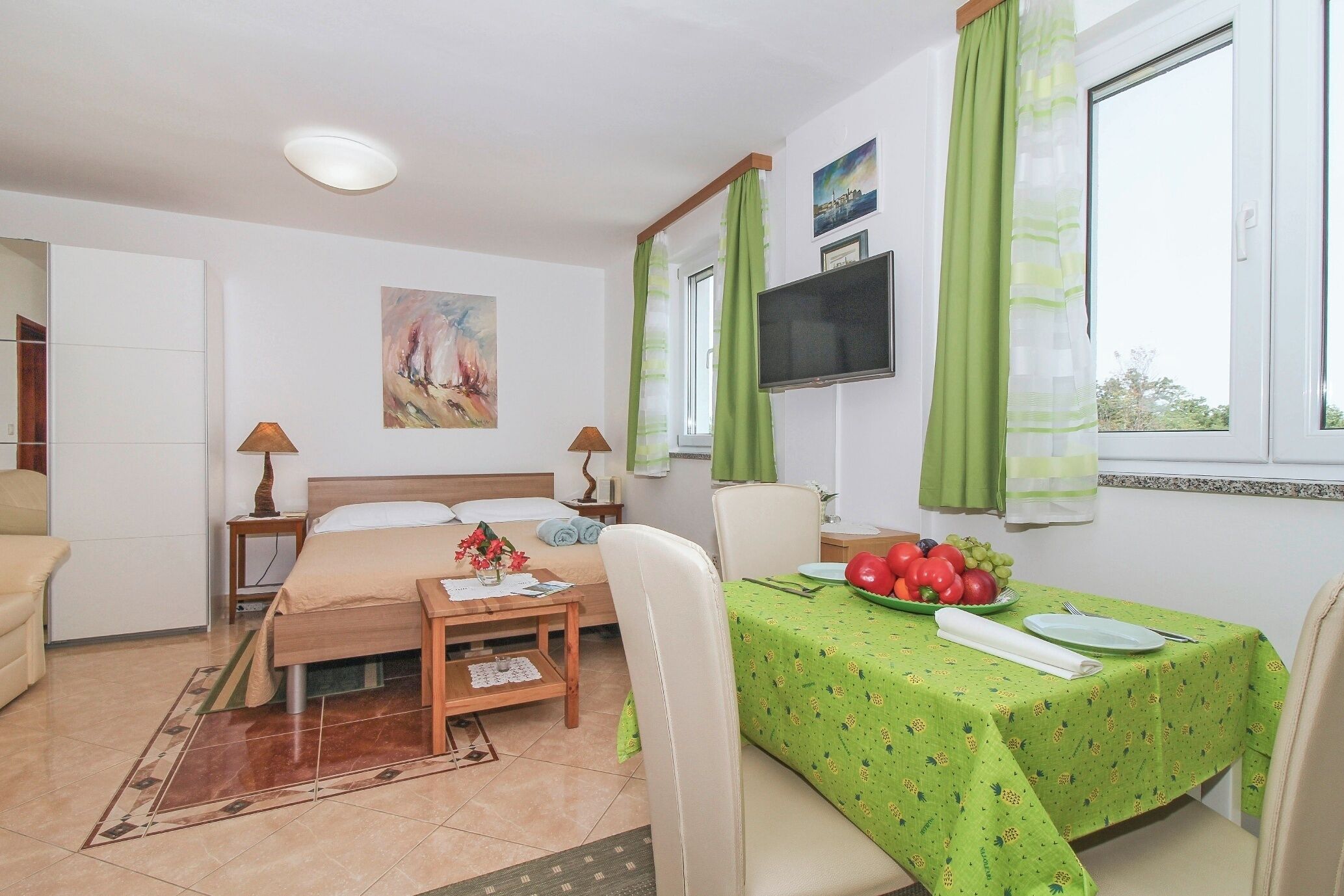 Une chambre d’hôtel avec un lit, un bureau et une chaise, une table à manger recouverte d’une nappe verte et un téléviseur fixé au mur.