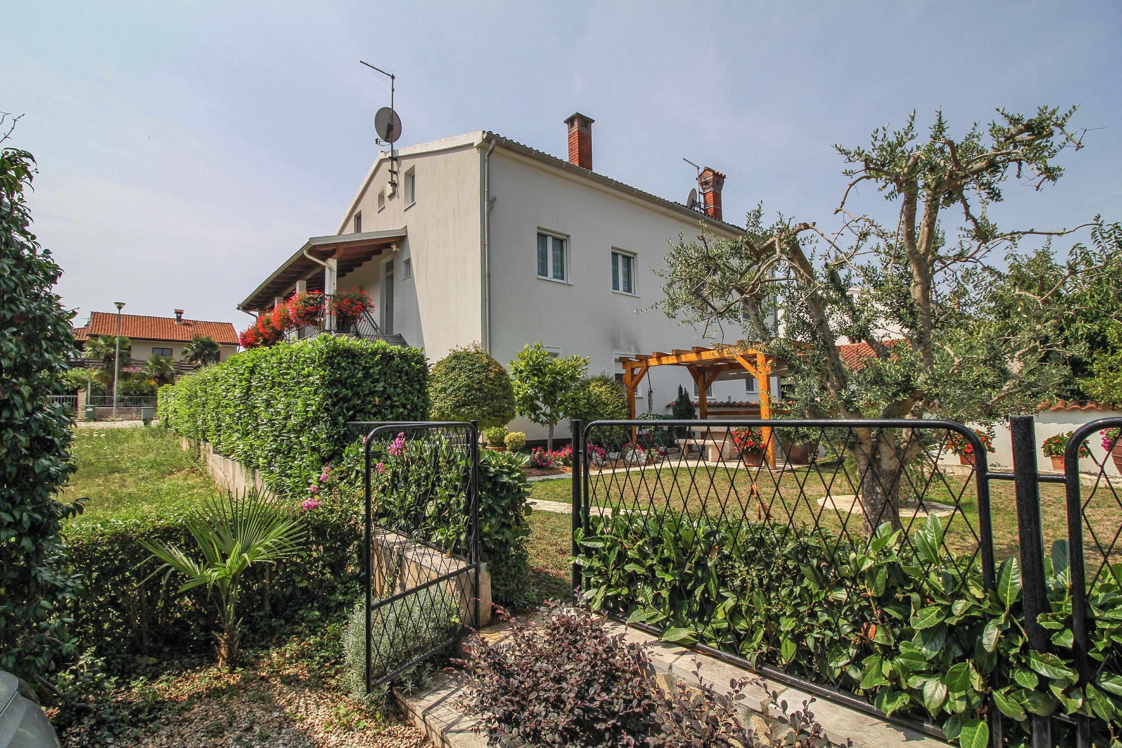 A two-story house with a balcony, surrounded by a garden with various plants and a metal fence.