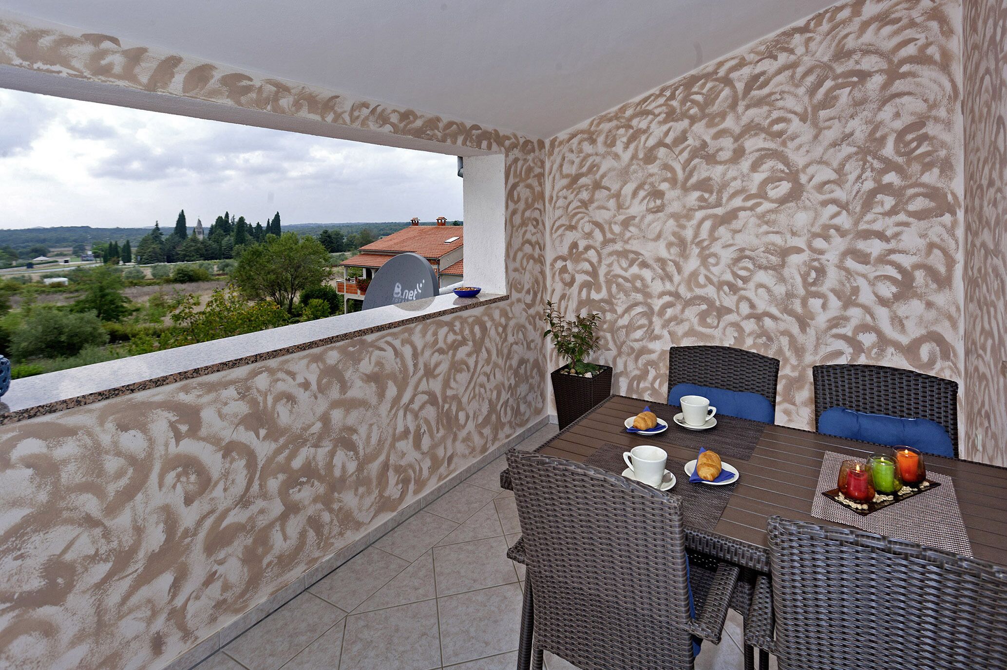Un balcón con una mesa de comedor para cuatro personas, sillas de mimbre, una planta en maceta y vistas a un paisaje distante.