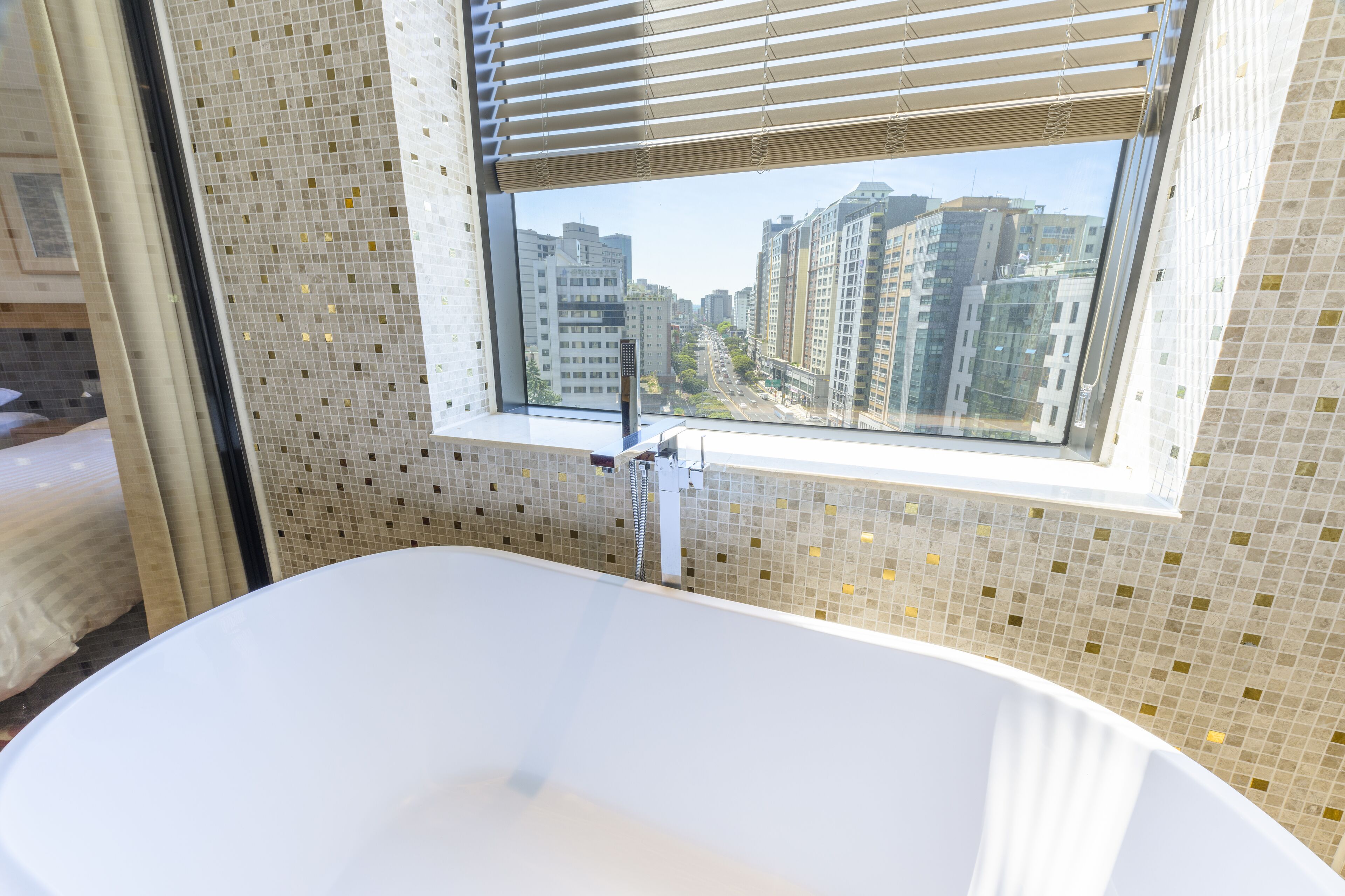 A modern bathroom with a large bathtub, a window with a city view, and mosaic tile walls.