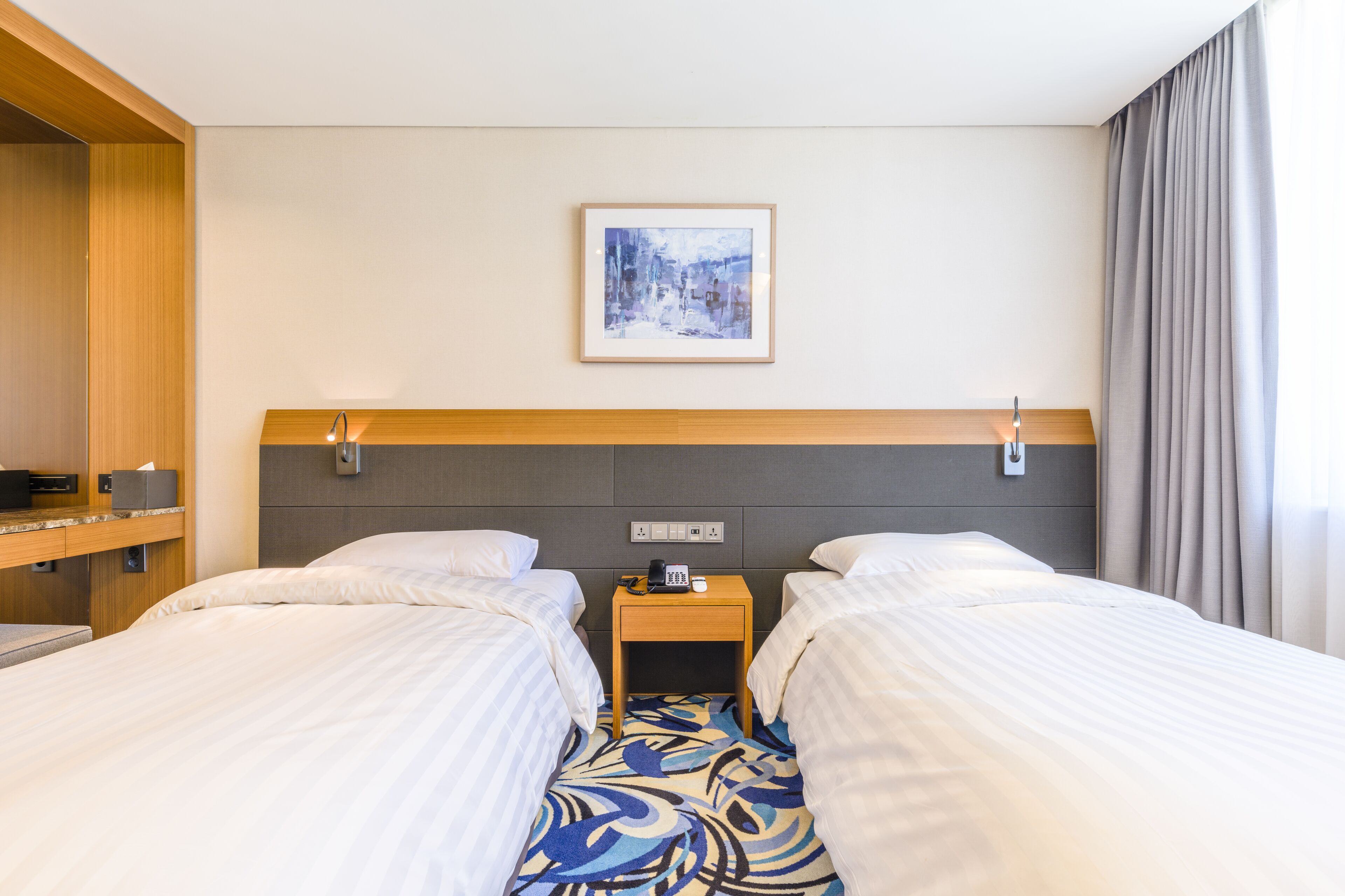 Habitación de hotel con dos camas, una mesita de noche con lámpara, un cuadro en la pared y una ventana con cortinas.