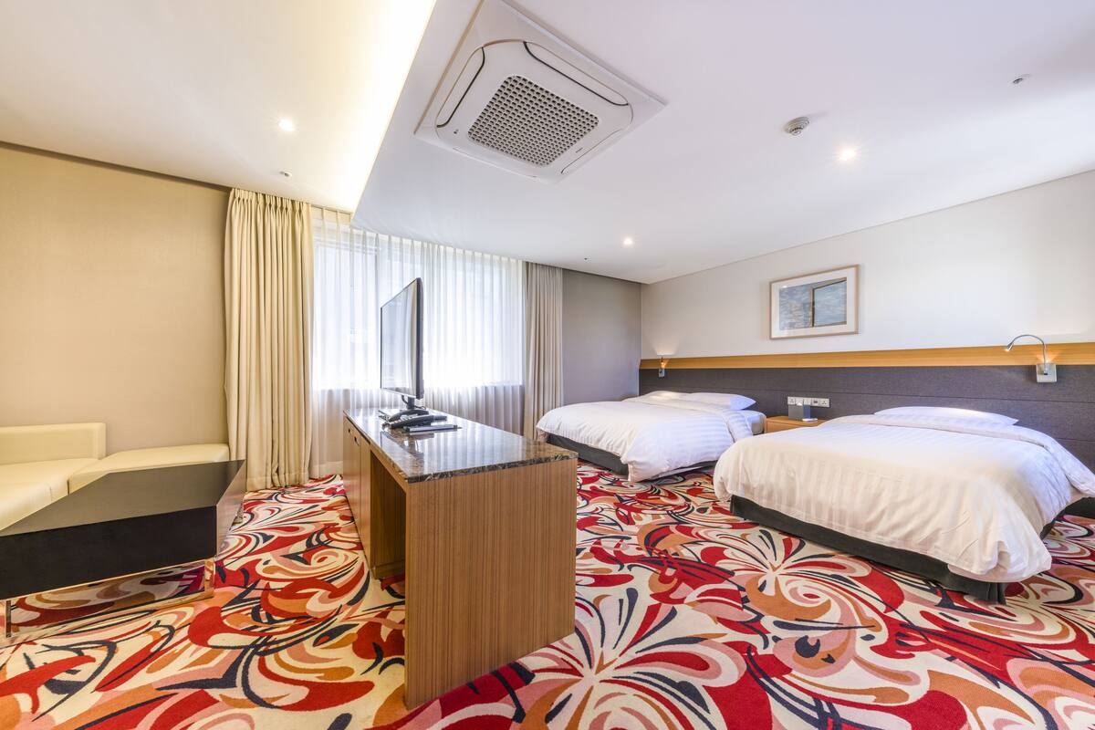 Ein Hotelzimmer mit Schreibtisch, zwei Betten, einem großen Fenster mit Vorhängen und einem gemusterten Teppich.