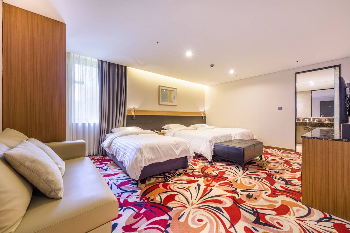 Ein Hotelzimmer mit einer Couch, zwei Betten, einem farbenfrohen Teppich, einer Wandpaneele aus Holz, einem Gemälde und einem Spiegel.