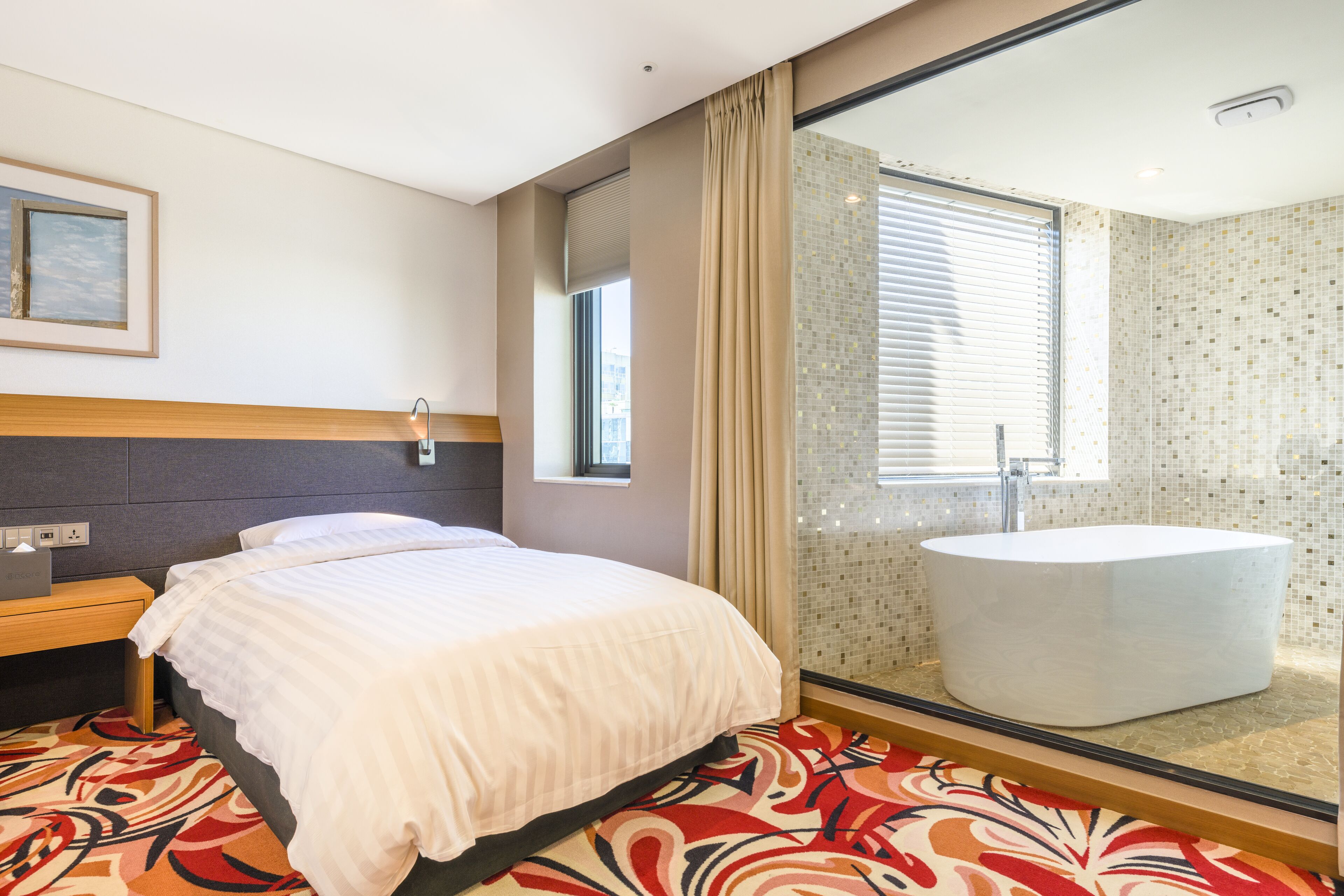 Una habitación de hotel moderna con una cama grande, un baño con bañera independiente y una ventana con persianas.