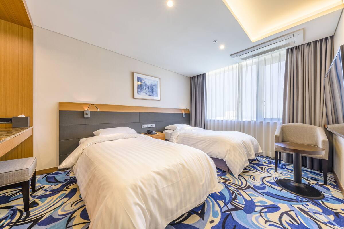 Ein Hotelzimmer mit zwei Betten, einem Schreibtisch, einem Stuhl und einem großen Fenster mit Vorhängen.