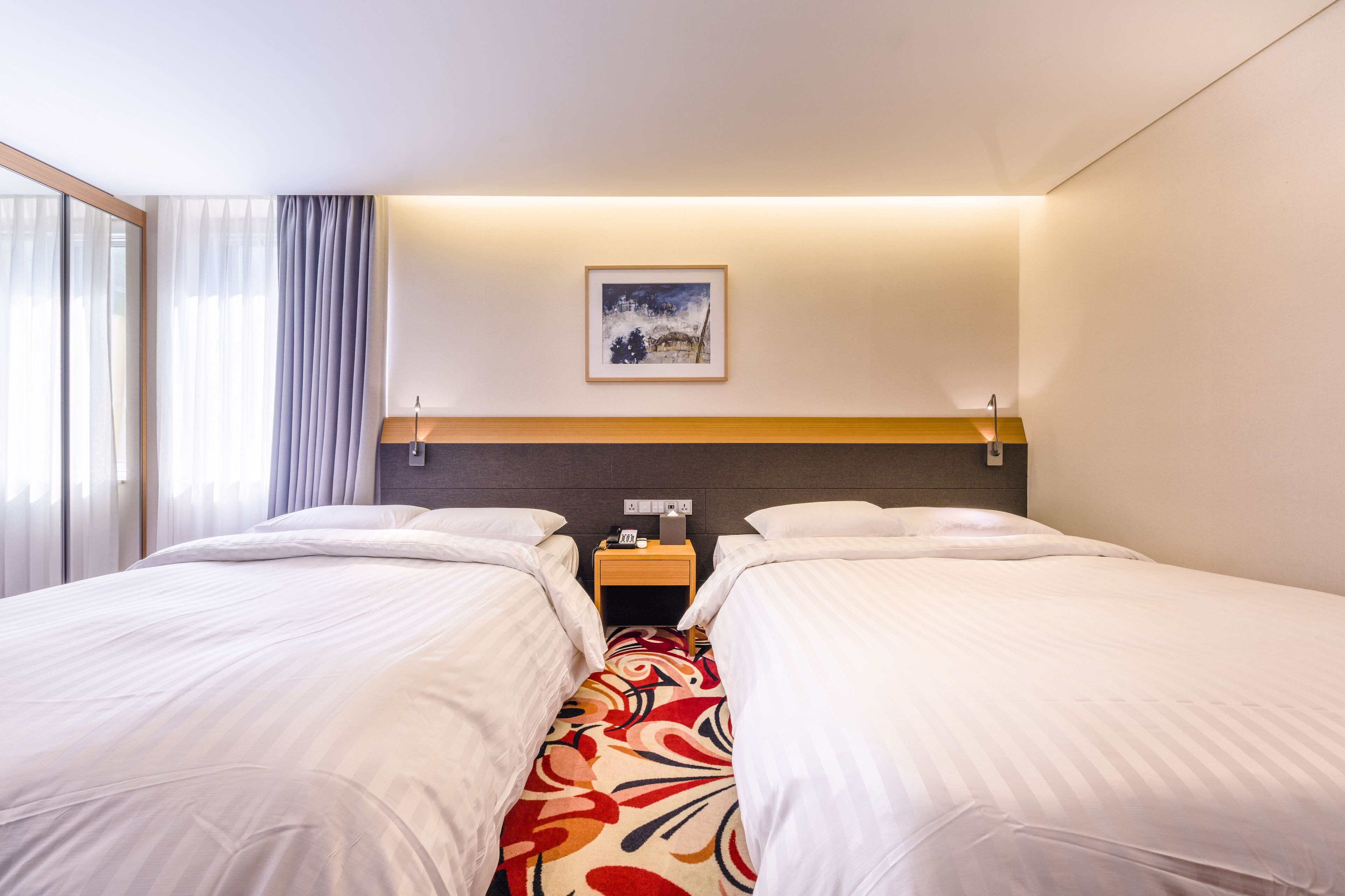 A hotel room with two beds, a nightstand, a framed picture on the wall, and a window with curtains.