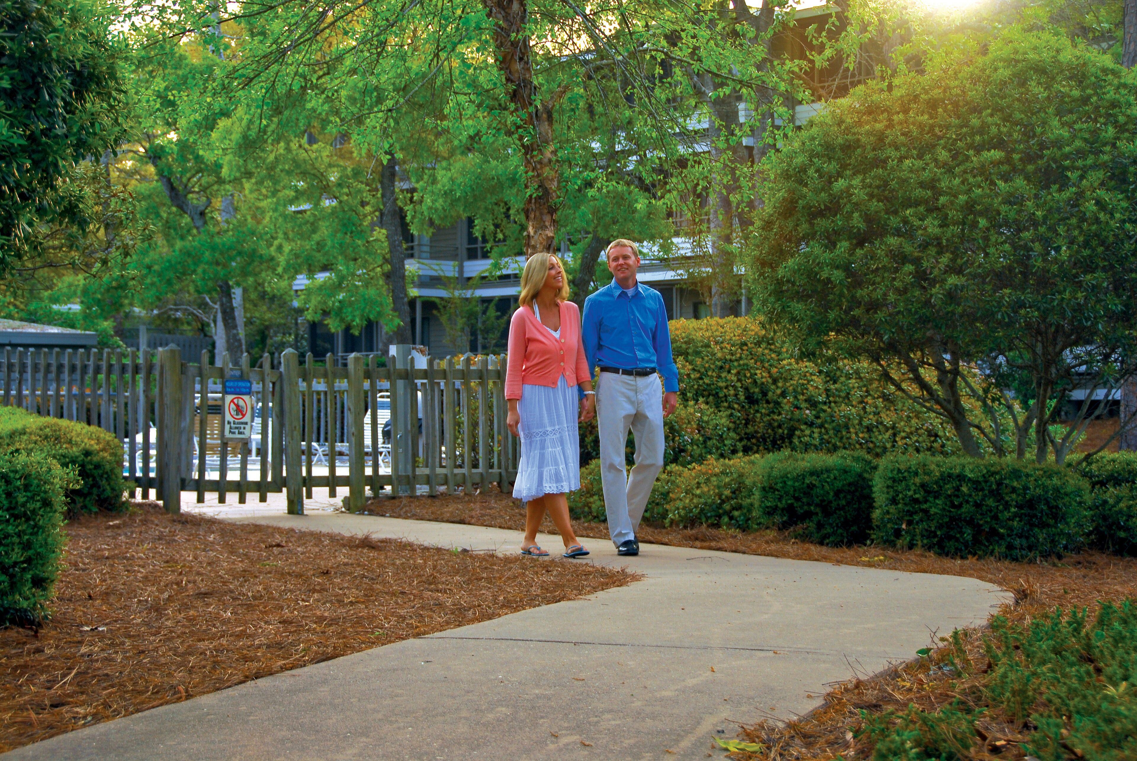 A couple walking on a sidewalk in a residential area with trees and a wooden fence.