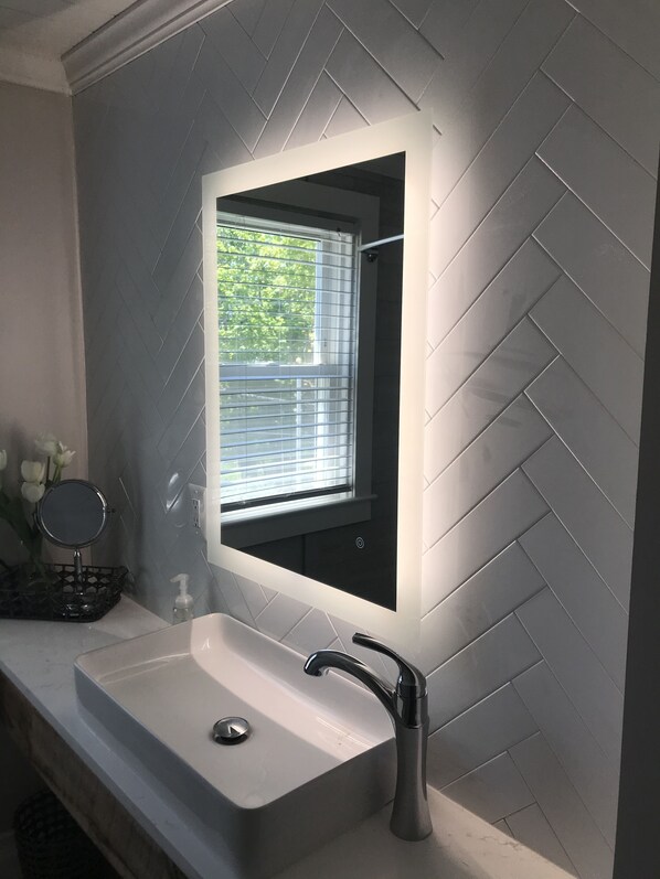 Un baño con lavamanos blanco, un espejo y una ventana con persianas.
