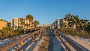 A wooden boardwalk leading to a beachfront property with multiple-story buildings, palm trees, and a clear sky.