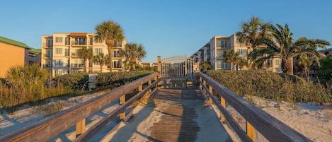 A wooden boardwalk leading to a beachfront property with multiple-story buildings, palm trees, and a clear sky.