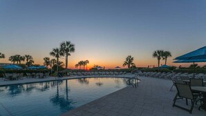 A swimming pool with lounge chairs and umbrellas, palm trees, and a sunset in the background.