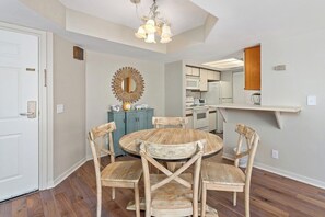 Uma sala de jantar com uma mesa de madeira redonda e quatro cadeiras, um espelho decorativo e uma cozinha ao fundo.
