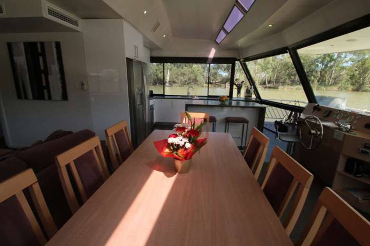 Un coin repas avec une table et des chaises en bois, un bouquet de fleurs et une vue sur l’eau grâce à de grandes fenêtres.