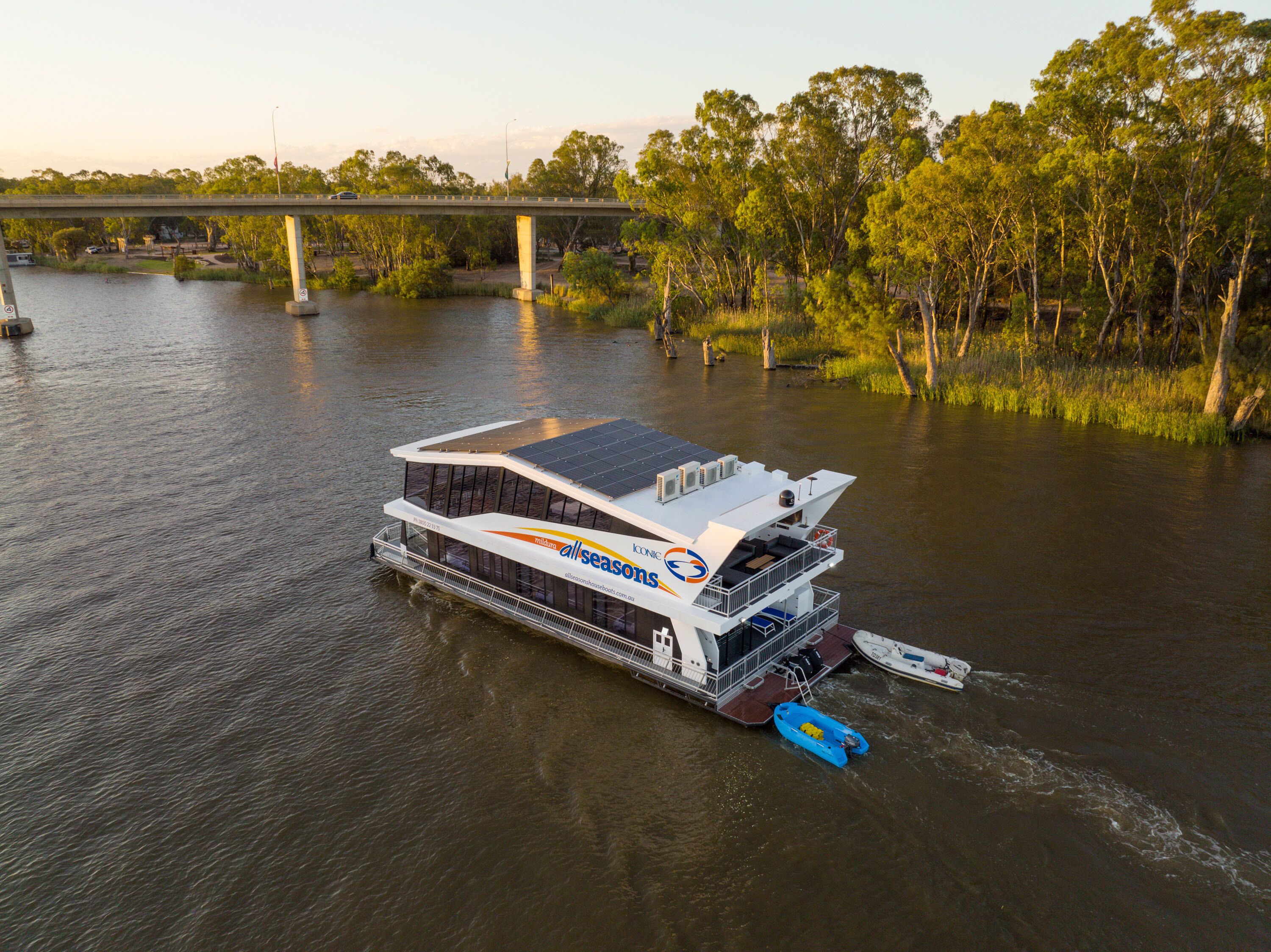 A riverboat with a covered deck and a small boat attached, docked at a river with a bridge in the background.
