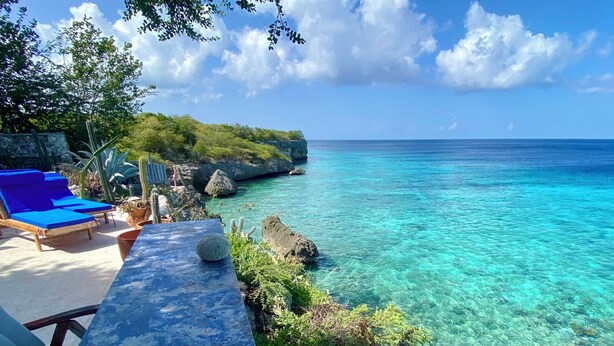 A clear view of a tropical beach with a rocky shoreline, a lounge area with blue chairs, and a table with a decorative item. - SEA FRONT-Private, natural, couples-only...unique paradise focused on peace (Banda Abou)