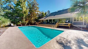 A rectangular swimming pool with a wooden deck and steps leading to a covered area with outdoor seating.