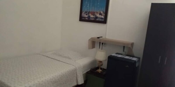 A small, neatly arranged room with a bed, a desk with a lamp, a small cabinet, and a wardrobe. - Lasa Residence (Oranjestad)