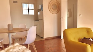 On aperçoit un coin repas avec une table ronde, des chaises blanches et un fauteuil jaune. Au fond, on distingue une cuisine équipée d’un four à micro-ondes et d’un réfrigérateur.