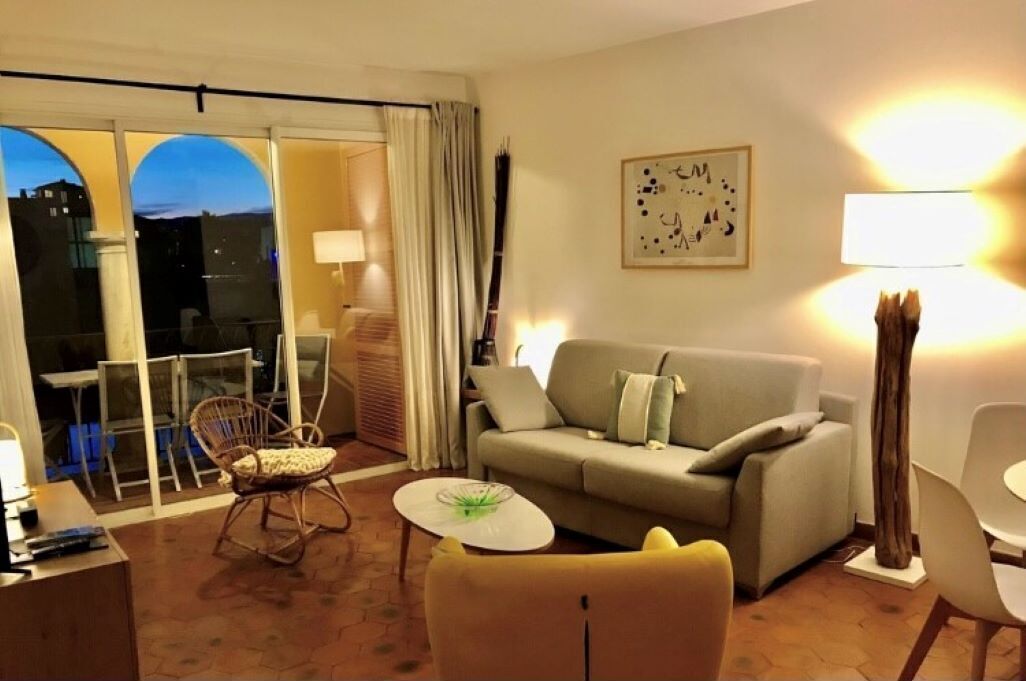 Una sala de estar con un sofá, una silla, una mesita y una lámpara. Hay una ventana con cortinas y vistas de edificios.
