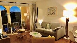 Un salon avec un canapé, un fauteuil, une petite table et une lampe. Il y a une fenêtre avec des rideaux et une vue sur les bâtiments à l'extérieur.