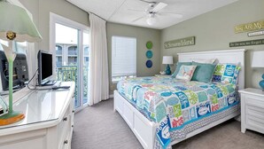 Ein Schlafzimmer mit einem Bett, einem Schreibtisch mit Computer und einem Deckenventilator, mit Blick auf den Balkon.