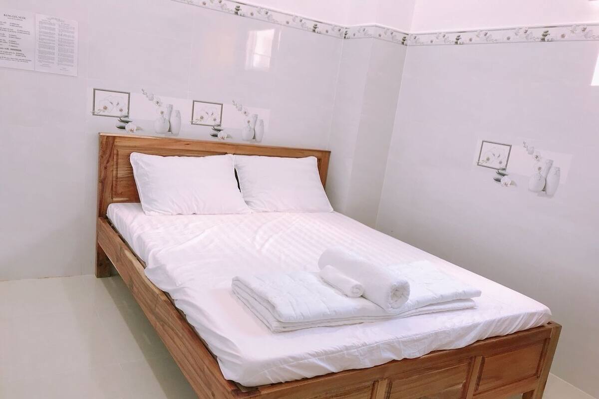 Un lit bien fait, avec du linge de lit blanc et une tête de lit en bois, dans une chambre propre et blanche, dont le sol est recouvert de carreaux.
