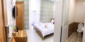 A hotel room with a bed, a wooden bedside table, a TV mounted on the wall, and a bathroom with a toilet and shower. - Thanh Vinh Hotel (Ho Chi Minh City)