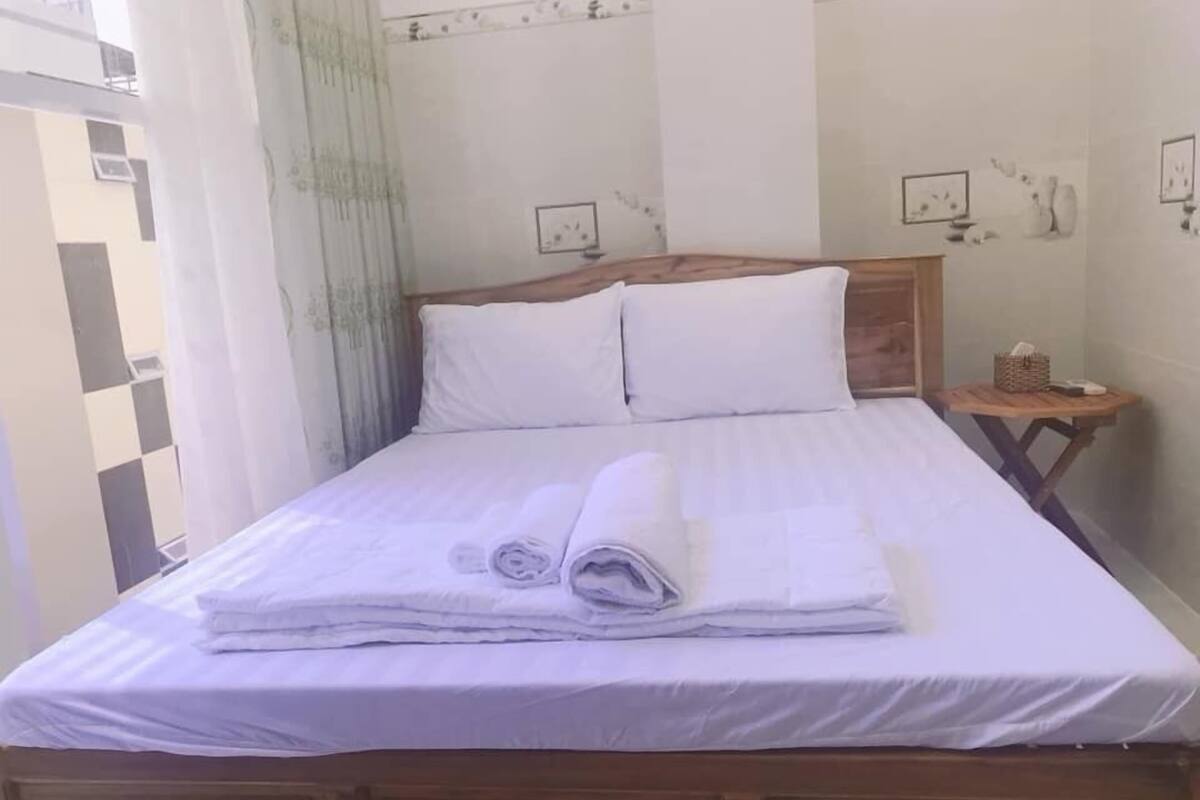 Un lit bien fait, avec du linge de lit blanc et des serviettes pliées, une tête de lit en bois et une petite table de chevet en bois sur laquelle se trouvent un panier et un téléphone.
