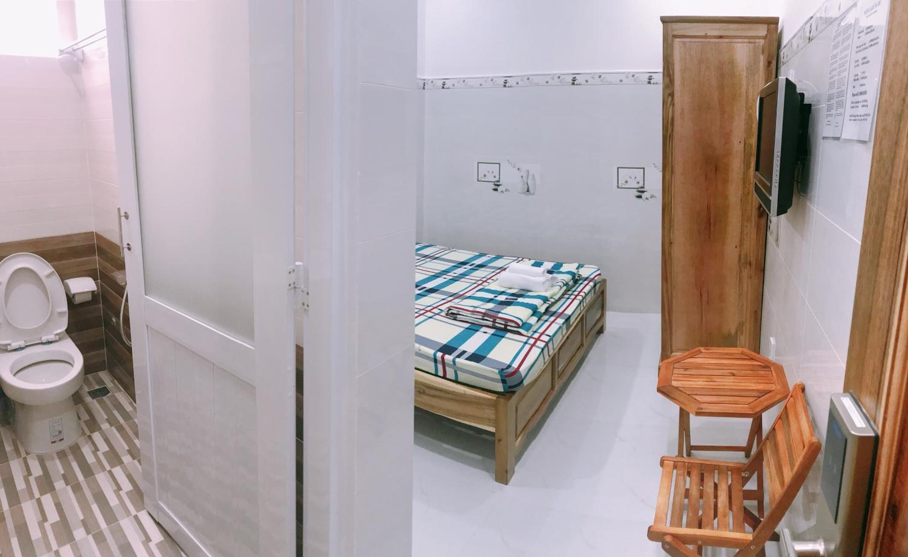 A small, clean room with a bed, a wooden chair, a wooden wardrobe, and a toilet.