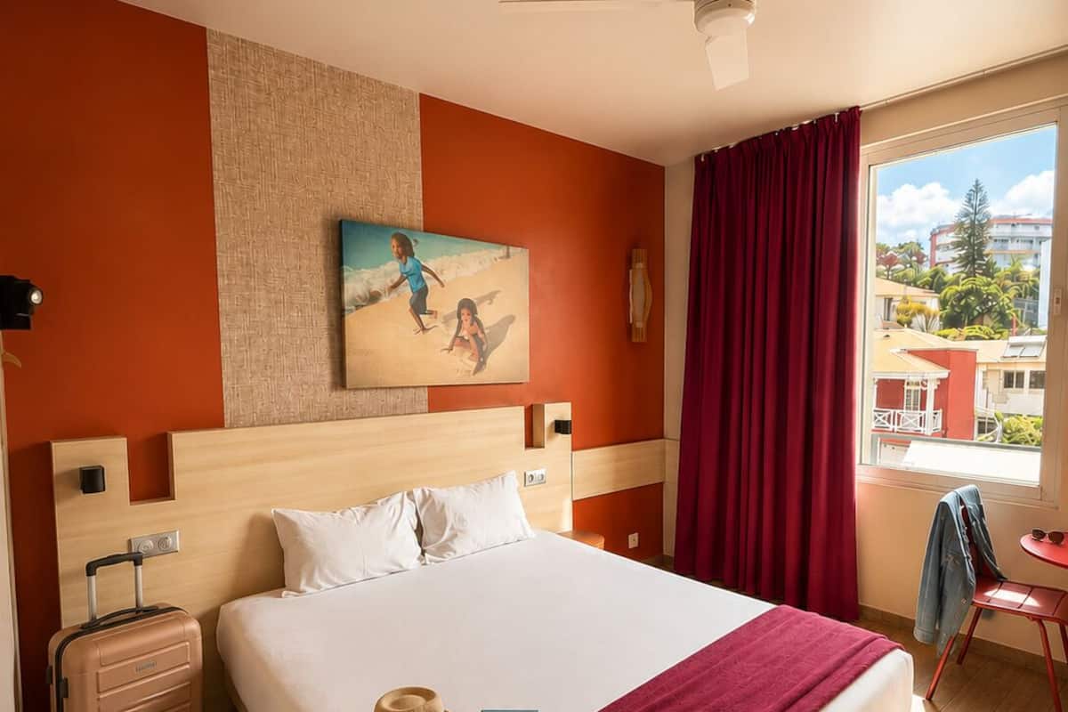 A hotel room with a bed, a suitcase, a hat, a book, a painting, a ceiling fan, and a view of buildings and trees.