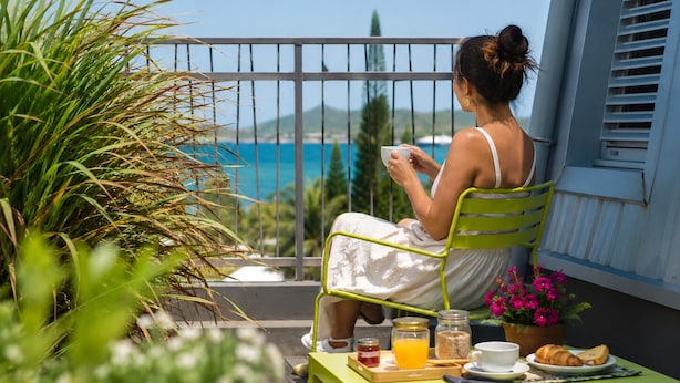 A woman sitting on a green chair on a balcony with a table in front of her, holding a cup. - Hotel Gondwana - City GREEN (Nouméa)