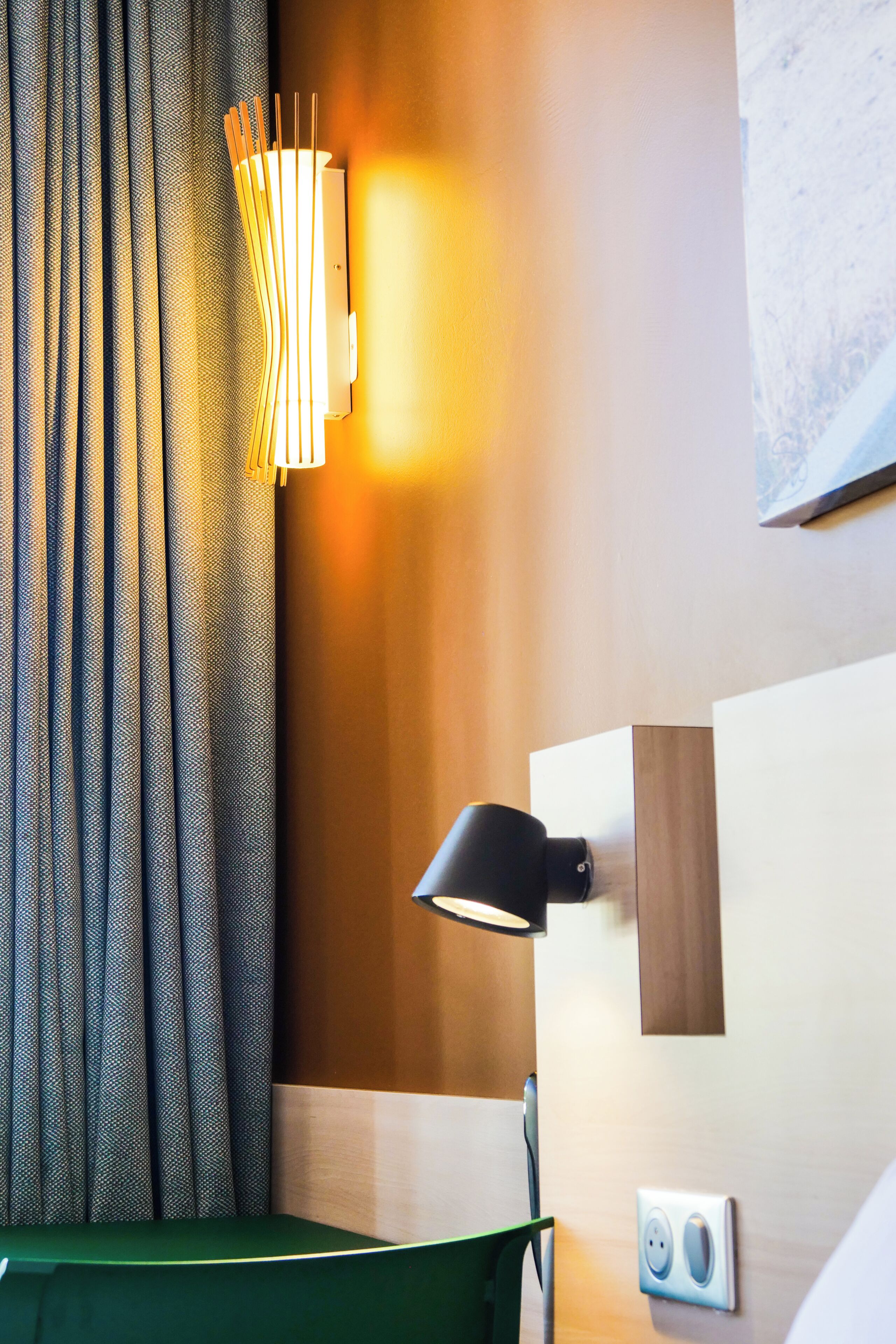 A hotel room with a bed, a wall-mounted lamp, a power outlet, and a curtain.