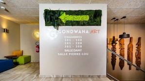 A lobby with a reception desk, green and blue seating, a staircase, and a wall displaying art pieces and a sign for 'Gondwana Art' with room numbers. - Hotel Gondwana - City GREEN (Nouméa)