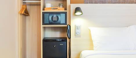 A hotel room with a bed, a nightstand, a desk with a lamp, a microwave, a safe, and a painting of a person by the sea.