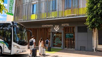 Entrada de un hotel con fachada moderna, un autobús estacionado adelante y personas con equipaje.