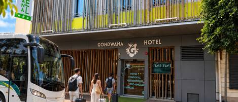 A hotel entrance with a modern facade, a bus parked in front, and people with luggage.