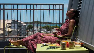Ein Balkon mit Frühstückstisch, eine Person sitzt darauf, Blick auf Gebäude und Wasser.